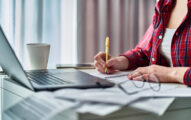 A person in a red plaid shirt writes in a notebook at a desk with a laptop, papers, and a coffee cup.