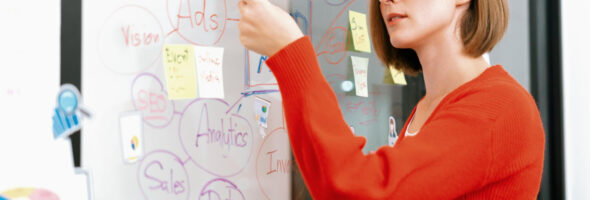 Woman in a red sweater writing on a glass board filled with marketing notes, diagrams, and sticky notes.